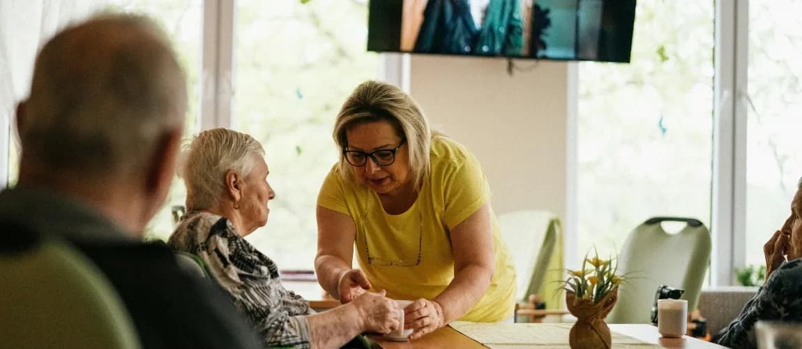 Eine Frau reicht einer Seniorin in einem Aufenthaltsraum eine Tasse.