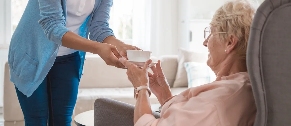 Eine Pflegeperson reicht einer älteren Dame einen Tee.