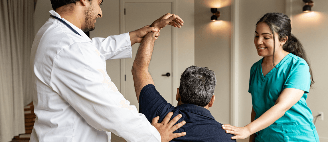 Ein Arzt hebt den Arm eines Patienten an, eine Frau legt ihm lächelnd eine Hand auf die Schulter.