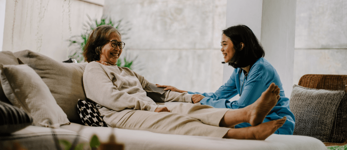 Eine ältere Frau liegt auf dem Sofa, daneben sitzt eine Pflegefachkraft, beide lächeln.