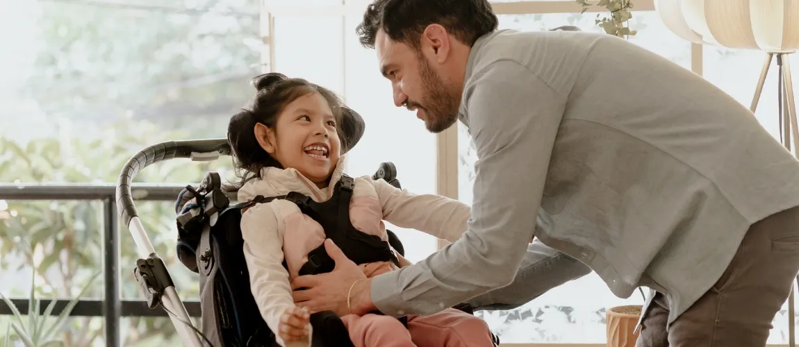Ein Mann positioniert ein lächelndes Mädchen in einen Rollstuhl.