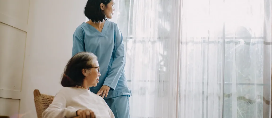 Eine Seniorin sitzt auf einem Stuhl, daneben steht eine Pflegekraft-beide blicken aus dem Fenster.