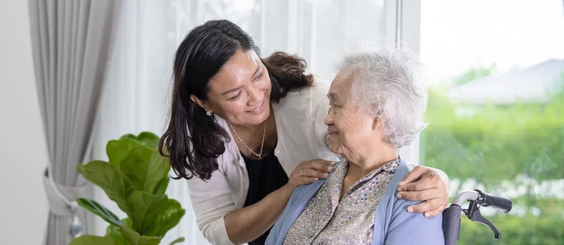 Eine Senior:in sitzt im Rollstuhl, eine Pflegeperson legt ihr die Hände auf die Schultern.