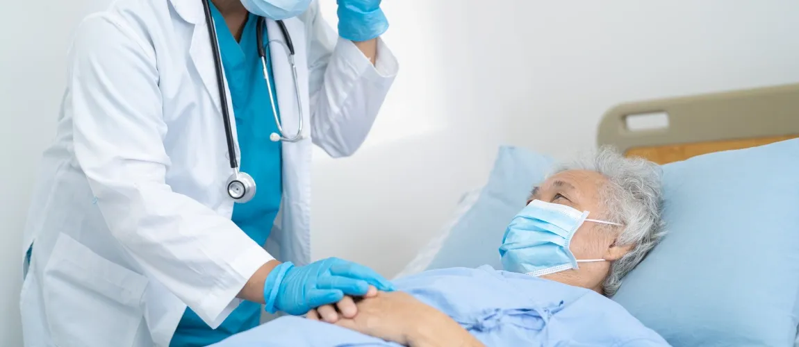 Eine Seniorin liegt im Bett, eine Pflegefachkraft steht daneben und legt ihr eine Hand auf.