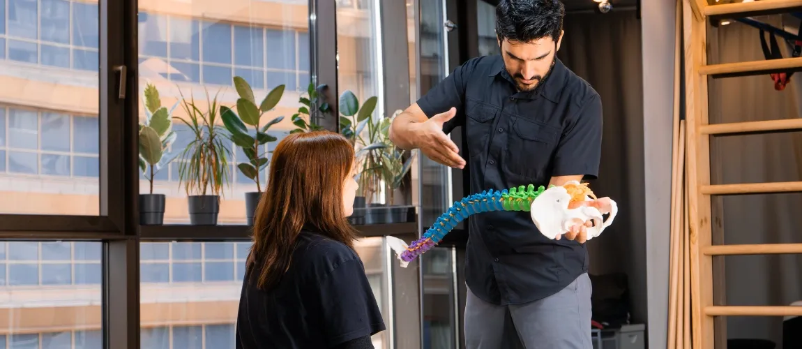 Ein Mann hält ein Modell der Wirbelsäule in der Hand und zeigt darauf, vor ihm sitzt eine Frau.