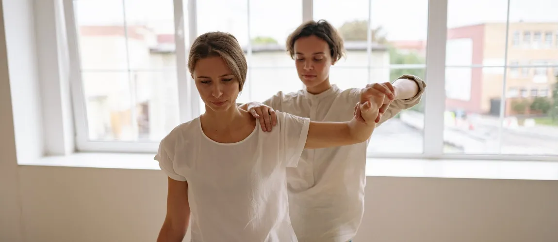 Eine Frau sitzt auf einer Liege, eine Therapeutin hebt ihren Arm an.