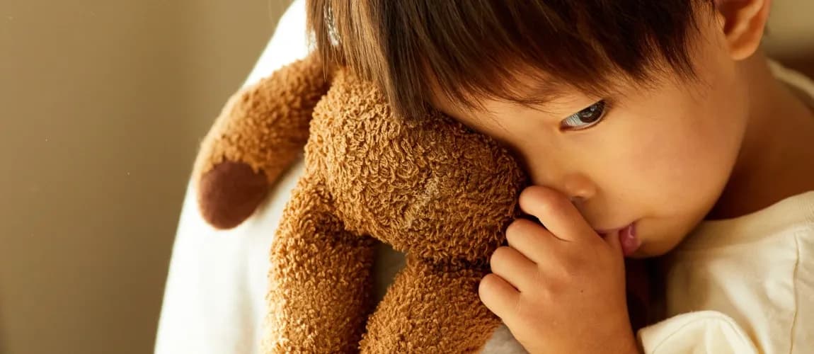 Ein Kleinkind umarmt einen Teddybären und lutscht am Daumen.
