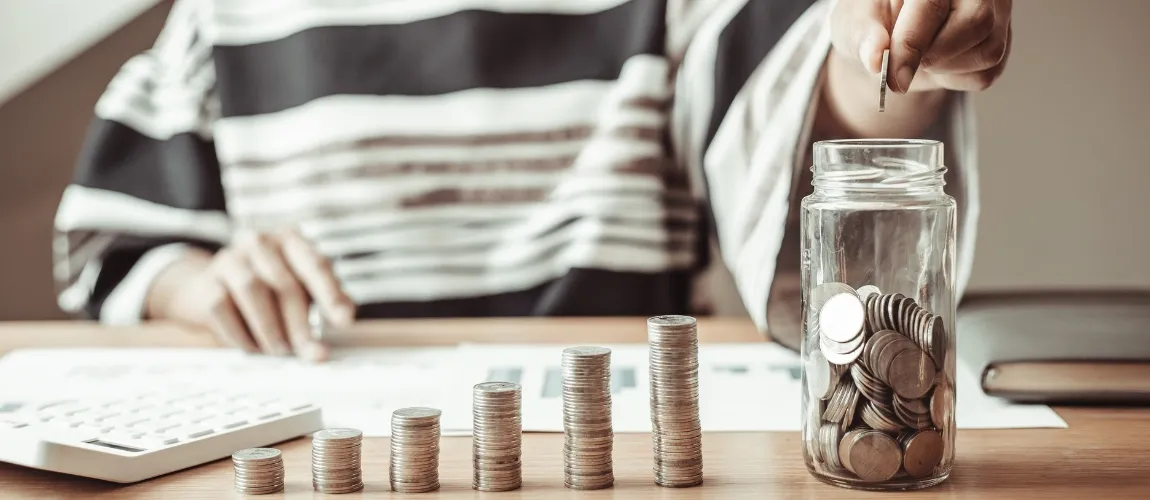 Eine Person sitzt an einem Schreibtisch mit Münzen und legt eine Münze in ein Glas.
