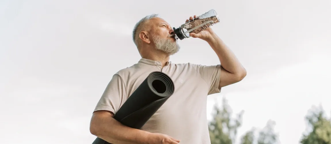 Ein Senior hält eine Sportmatte unter dem Arm und trinkt aus einer Flasche.