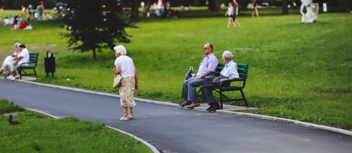 Eine Seniorin geht einen Weg im Park entlang, an der Seite sitzen Menschen auf Bänken.