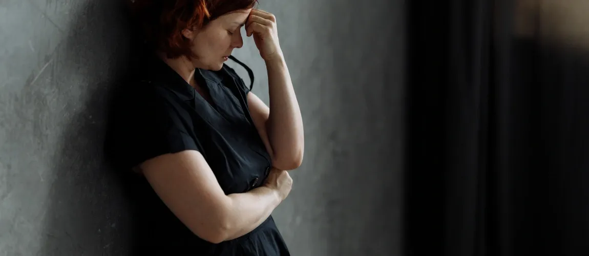 Eine Frau in schwarzer Kleidung lehnt sich an eine graue Wand, sie hält sich eine Hand an die Stirn.